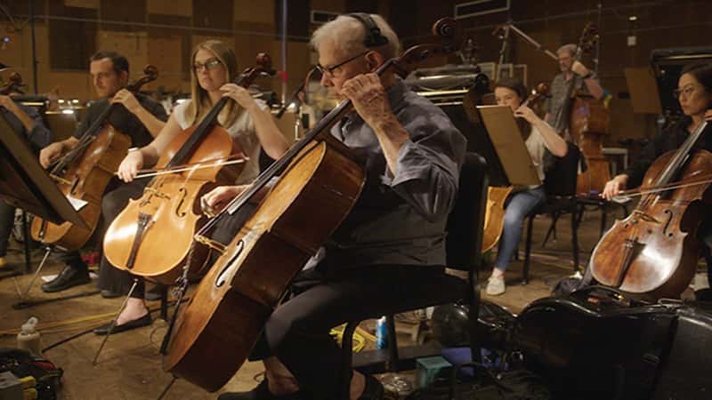 Several people playing cellos in a well-lit room, engaged in a collaborative music session.